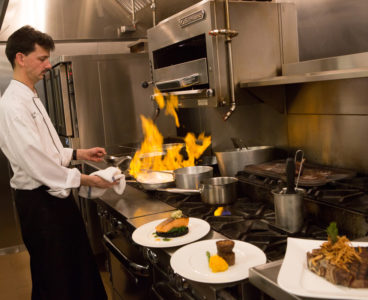 chef holding flaming pan over oven. plated food off to the side